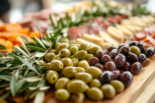 A close-up view of a charcuterie board featuring green and black olives, olive branches, assorted cheeses, sliced meats, and dried fruits arranged neatly